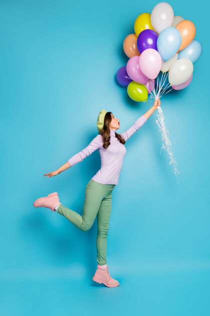 乐趣疯狂女士张嘴举着许多气球随风升起的全长垂直照片穿紫色套头衫贝雷帽绿色长裤靴子孤立的蓝色墙壁裤子简介飞行