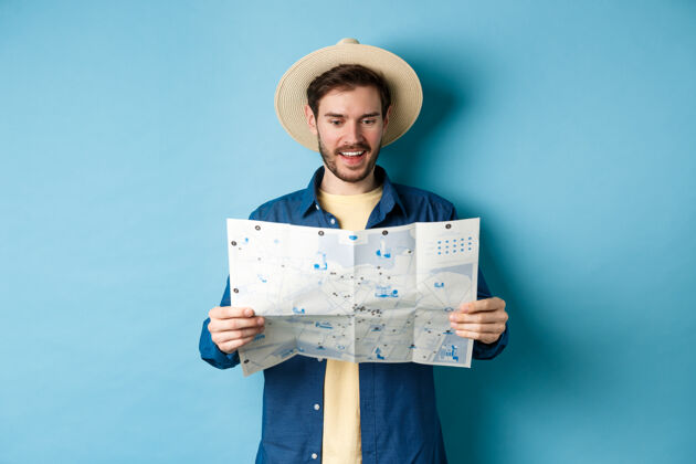 帽子兴高采烈的游客在夏日帽子上规划旅游线路度假 望着叹气看地图 微笑着兴奋 站在蓝色的背景上地图帅气阅读