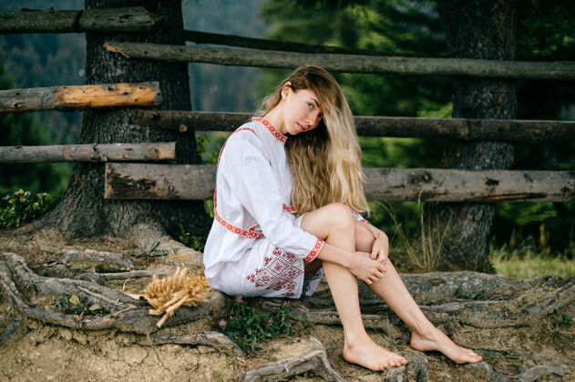 赤脚年轻迷人的赤脚金发女孩 穿着白色连衣裙 戴着饰物 坐在户外的木栅栏旁篱笆金发衣服