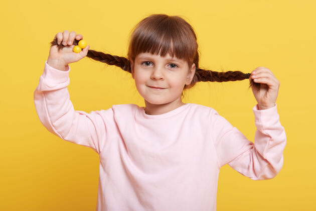 孩子微笑的女孩手挽着辫子 直视着黄色背景下的镜头 迷人的女孩儿独自玩耍 穿着淡粉色休闲衬衫的孩子乐趣毛衣情感