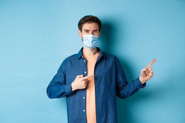 男人Covid和医疗保健概念戴着医学面具的帅哥指着旁边的空地 站在蓝色的背景上成人帅哥年轻
