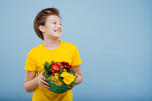 花快乐的学龄前男孩手里拿着花篮 穿着黄色t恤隔离在蓝色的墙上 复制空间 侧面视图玫瑰未成年男孩