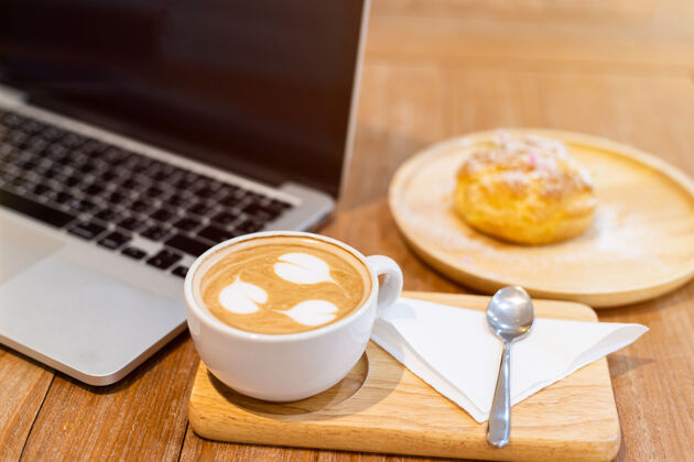 泡沫特写镜头与咖啡杯工作与笔记本电脑和自制profiteroles与choux奶油咖啡店一样的背景现代拿铁木材