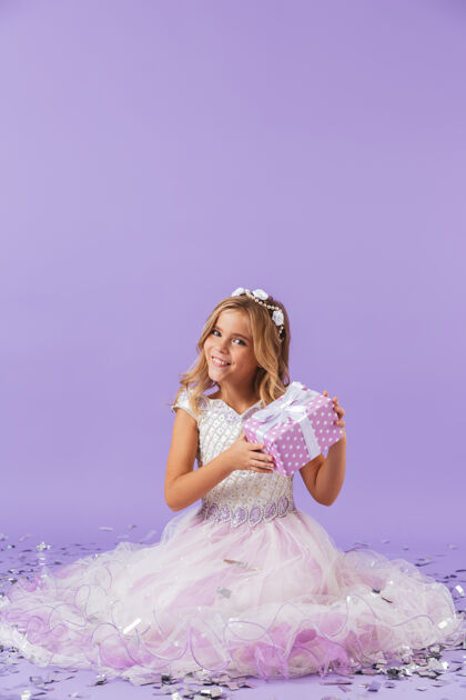 庆祝穿着公主裙的漂亮小女孩孤零零地坐在紫罗兰色的墙上 手里拿着礼物盒年轻爱美丽