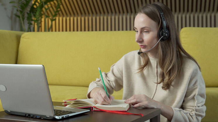 耳机戴着耳机的女人坐在家里的黄色沙发上 在笔记本电脑上视频聊天工作讨论在线