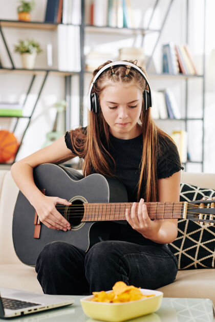 音乐天才创意少女戴着耳机坐在沙发上打开笔记本电脑前弹吉他技术声音微笑