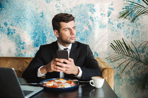职业一个英俊的年轻商人坐在咖啡馆里用笔记本电脑吃早餐或晚餐用手机吃饭的画面成功人士网络环境
