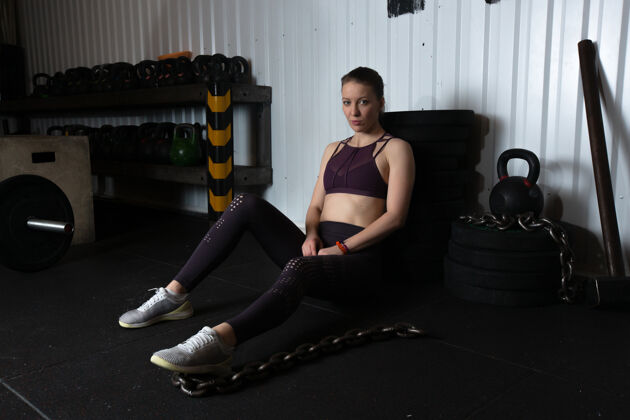 吸引力一位身穿紫色运动上衣和灰色紧身裤的年轻女运动员坐在运动器材附近的体育馆的地板上训练锻炼女性