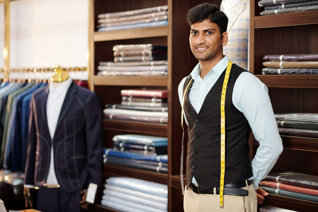 纺织业英俊微笑的印度裁缝的肖像 脖子上缠着卷尺 站在工作室里商人业主服装店