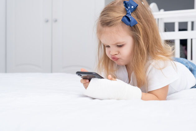 医学手拿石膏的小女孩躺在床上用智能手机 看动画片或教育视频石膏健康移动