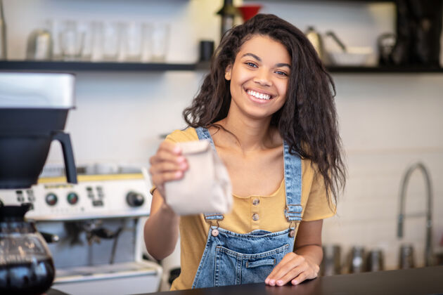 忙碌出售 咖啡开朗微笑着的年轻女子穿着牛仔工作服站在咖啡店柜台后面 手里拿着纸袋咖啡馆女人小企业