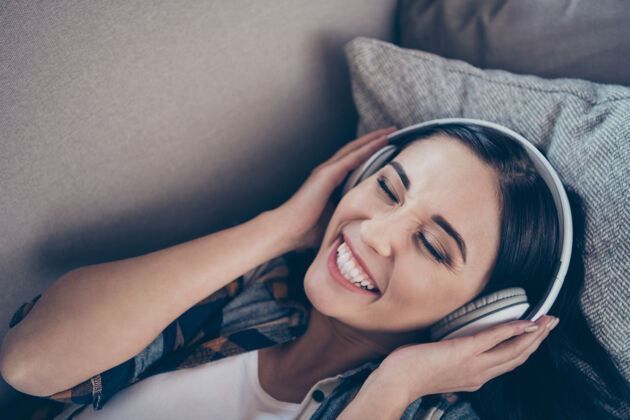 情人高角度观看照片惊人的女士在兴高采烈地听着最喜爱的收音机调频在现代耳罩躺在舒适的沙发上穿着休闲服公寓室内有趣耳机欢乐