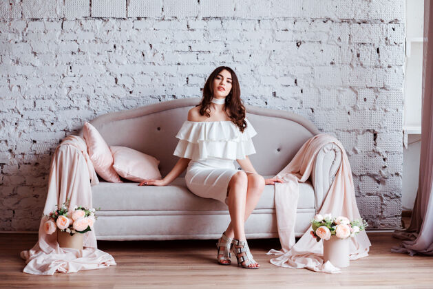 时尚身着白色连衣裙的迷人女子正坐在装饰着粉色枕头的沙发上姿势新娘枕头