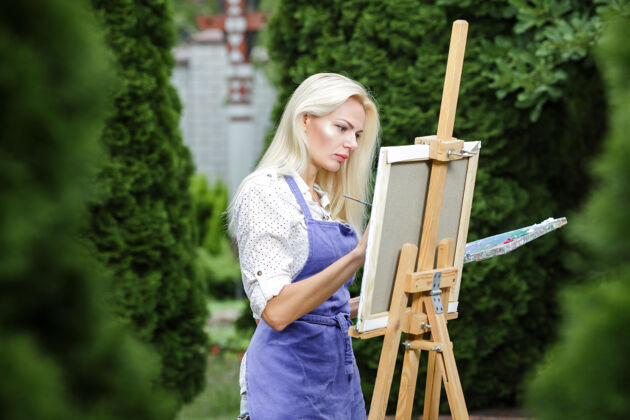 女性美丽的金发女画家手里拿着画笔在花园里的画布上画画户外水人物