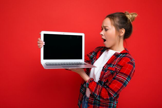 数字照片中的年轻女子手持手提电脑 身穿红色衬衫 看着红色屏幕上孤立的显示器背景.mock起来显示器技术上网本