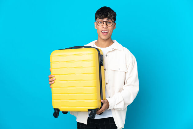 年轻年轻人在度假时带着旅行箱和蓝色的背景感到惊讶简短行李旅行
