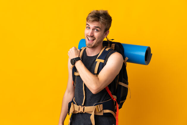 帅哥一个年轻的登山者背着一个大背包在黄色的背景上庆祝胜利男人运动旅游者
