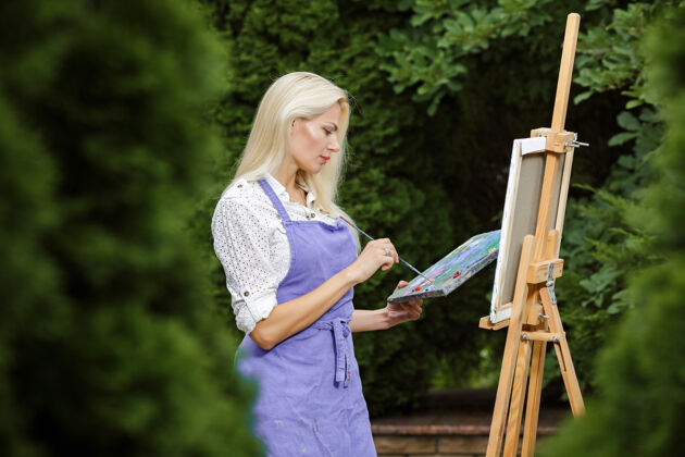 白色美丽的金发女画家手里拿着画笔在花园里的画布上画画图片年轻彩色