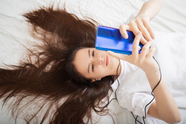满意一个女孩在家里戴着智能手机 戴着耳机听音乐 看手机 躺在床上生活方式女人心情