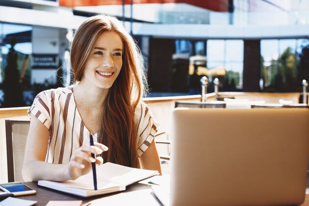 社交媒体年轻可爱的红发女影星坐在咖啡店外微笑着在笔记本电脑上做视频笔记本微笑手机