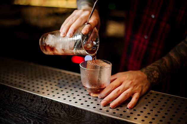 玻璃酒吧男招待用一个大冰块把新鲜而浓烈的威士忌鸡尾酒倒进玻璃杯里倒酒新鲜柜台