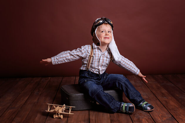 头盔一个戴着飞行员头盔的小男孩和一个木头玩具玩飞机男孩穿着格子衬衫和吊带灵感航空苍蝇