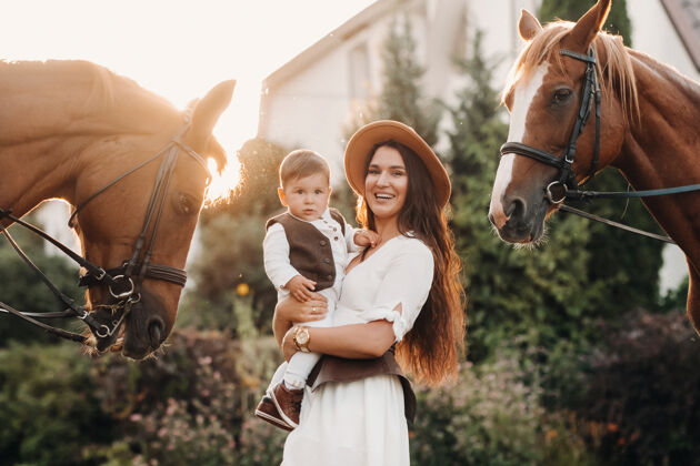 户外一位戴着帽子的母亲抱着儿子站在大自然中两匹美丽的马旁边一个有孩子的家庭被拍到和马在一起日时尚家庭
