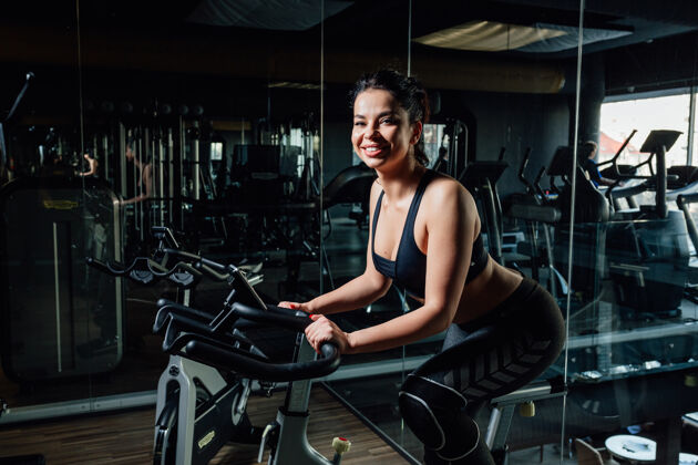 锻炼穿着运动服的迷人年轻女士微笑着坐在时尚健身房镜子旁的摩登健身车上女子骑物理