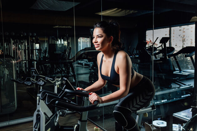 运动穿着运动服的迷人年轻女士微笑着坐在时尚健身房镜子旁的摩登健身车上健身女性现代