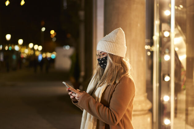 应用程序年轻的金发女郎戴着面具在商店橱窗前写着信息 在一个城市的夜晚 灯火通明背景.冬季气氛人户外连接