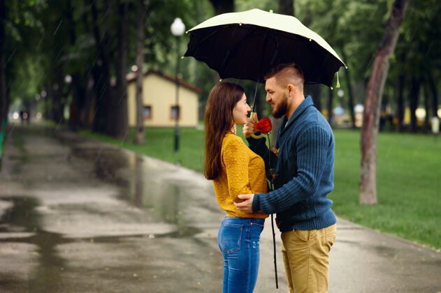 约会雨中夏日公园雨伞拥抱的情侣戴曼女人带着玫瑰在雨中走在小路上 雨天在巷子里男人女性丈夫
