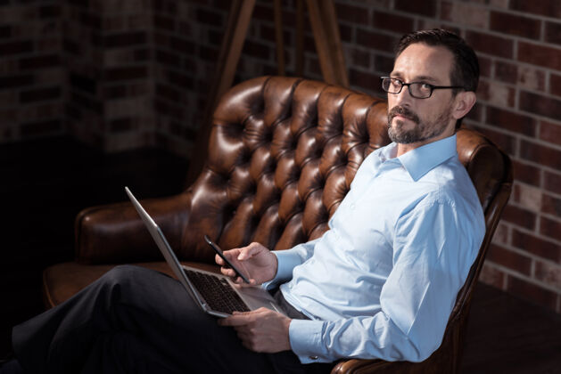 视力现代技术不错英俊的胡须男子坐在沙发上 看着他的手机 而他的膝盖上有一台笔记本电脑昂贵沙发设备