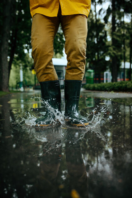 风暴男人穿着雨衣和胶靴在水坑里跳跃 天气潮湿胡同男人在夏季公园里摆姿势 下雨日.水保护 滴滴人家伙年轻
