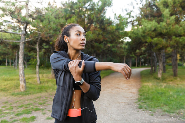 吸引力一幅20多岁的美籍非洲妇女穿着黑色运动服在绿色公园锻炼身体的照片美国保健耳机