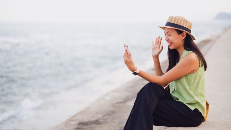 微笑在海边的海滩上 一位亚洲旅行者正在使用智能手机在线视频通话帽子旅游快乐