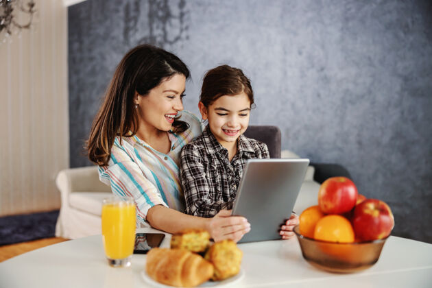 使用微笑着的母女俩早上坐在餐桌旁用平板电脑快乐家人在平板电脑上看动画片大笑沙发放松