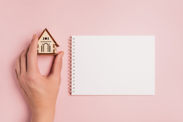 木材手持式房屋模型 粉色背景上有空白笔记本背景.投资 房地产 住宅 住房购买移动房地产