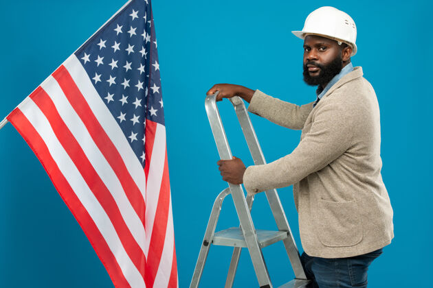 高加索种族年轻的留着胡须的非洲裔美国工头戴着防护头盔 穿着正装站在蓝色的美国国旗旁的梯子上选票非洲种族彩色形象