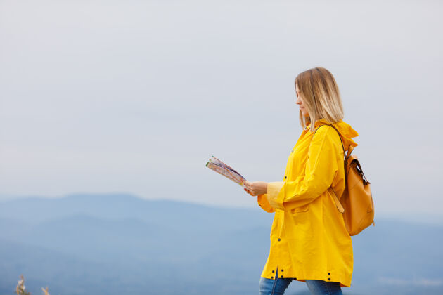 旅游穿着黄色雨衣的时髦女孩背着背包在山顶上看地图年轻绿色领导