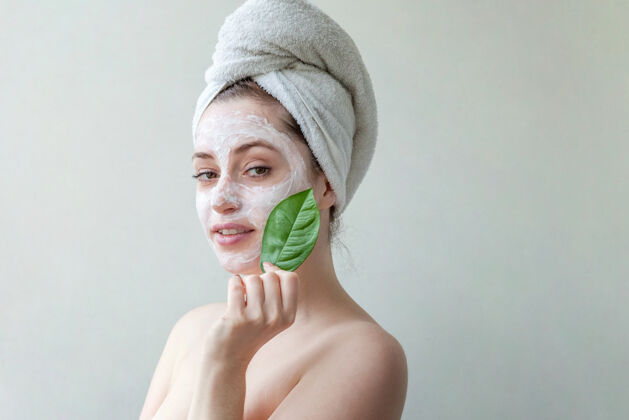 皮肤头戴毛巾 脸上涂有白色滋养面膜或乳霜 手拿绿叶 白色背景的美女肖像过程护理应用