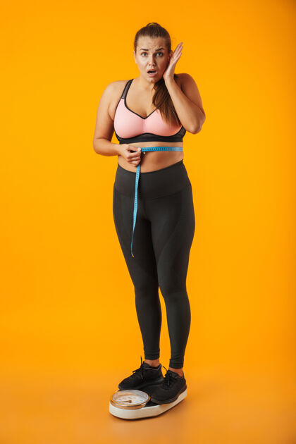 表情一个心烦意乱的超重的年轻女子穿着运动服站在黄色墙壁上 腰间拿着卷尺的全长肖像减肥运动体重秤