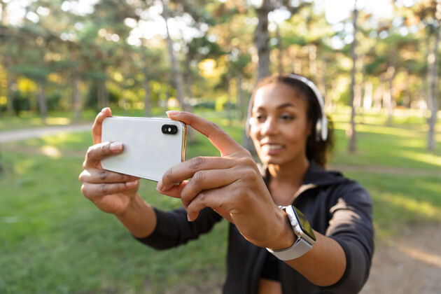 黑发20多岁的女运动员穿着黑色运动服 戴着耳机 在绿色公园散步时用手机自拍保健运动员运动