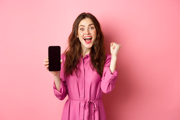 乐观科技概念兴奋的女孩展示手机屏幕 带着喜悦和幸福尖叫 在线赢钱 在智能手机应用程序上实现每日目标 粉红墙互联网聊天惊人
