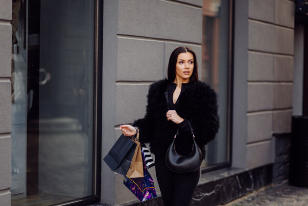 购物者一个棕色头发的女人穿着黑色的衣服 手里拿着五颜六色的有图案的购物袋 在一次成功的购物狂欢中走在外面 她享受着一天的温暖购物女士城市