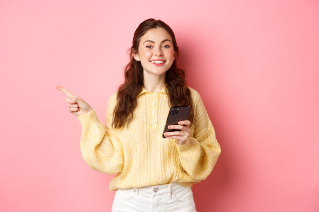 显示漂亮的现代女孩拿着智能手机 手指放在一边 一边用手机一边在版面上展示宣传文字 站在粉色的墙上请休闲情绪