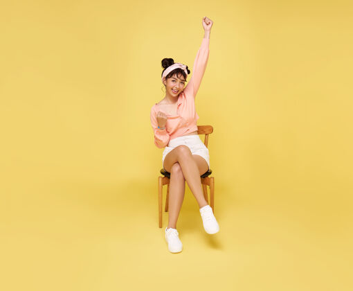 手臂快乐美丽的亚洲少女坐在椅子上 举手庆祝黄色墙壁上的手势兴奋的赢家成功概念微笑激情兴奋