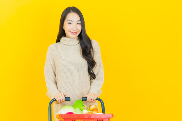 人一位美丽的亚洲年轻女子微笑着 黄色的墙上挂着超市的购物篮亚洲微笑消费主义