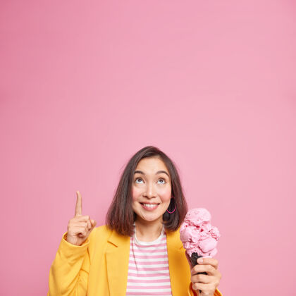 时尚时尚的千禧一代女孩 黑色短发点在头顶空白的复印件上 在粉色的墙上展示一些东西 吃着美味的树莓冰淇淋 含有很多热量季节装束姿势