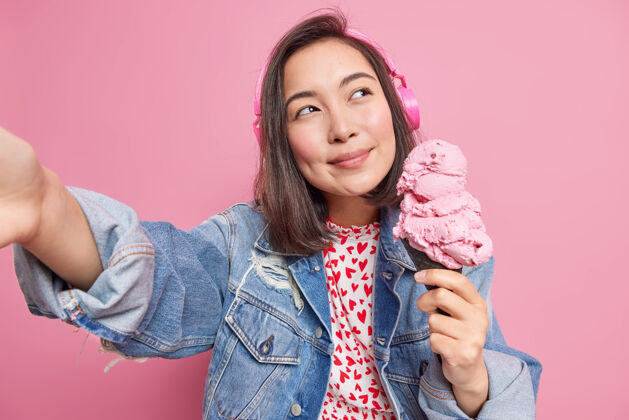 華夫餅干夢幻般的體貼美麗的深色頭發的亞洲女孩在華夫餅里擺出美味的大冰淇淋想象一些令人愉快的事情讓自己的照片通過立體聲耳機聽音樂有暑假休閑亞洲人姿勢