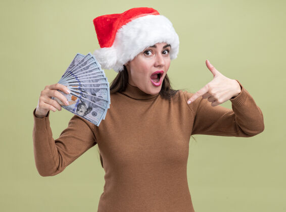 惊喜惊讶的年轻漂亮女孩戴着圣诞帽 手持现金指着隔离在橄榄绿墙上帽子点现金